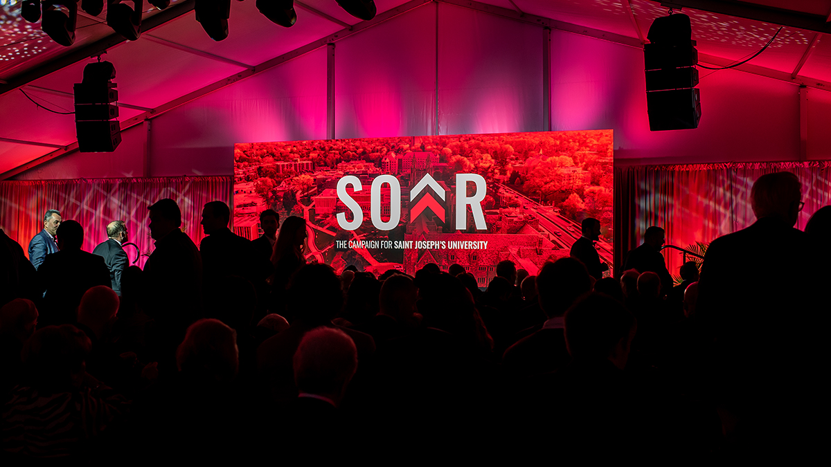 An audience waits inside a tent, crimson light illuminates a banner on stage that says SOAR