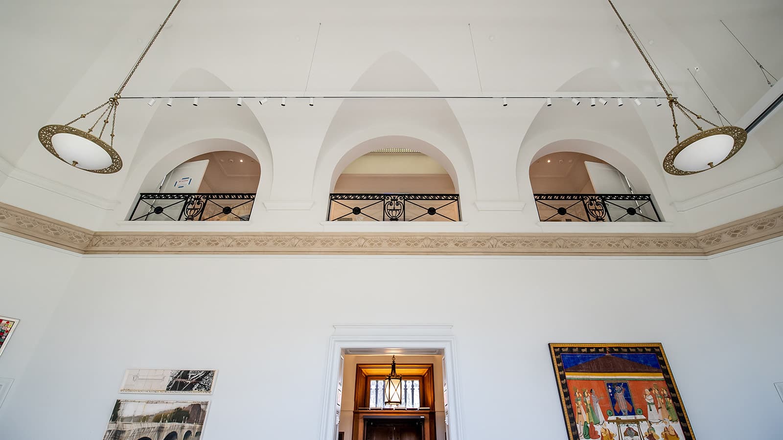 Angled photo of light fixtures in the Maguire Museum with frames on the wall and upper level arches behind